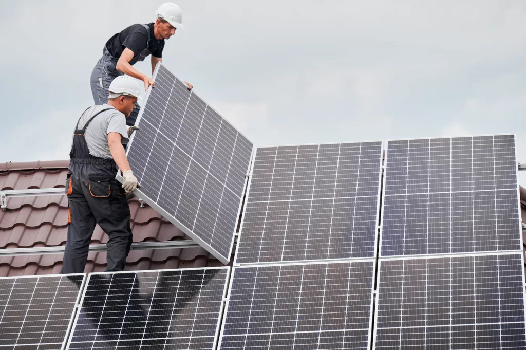 Technicians mounting photovoltaic solar moduls on roof of house. Builders in helmets installing solar panel system outdoors. Concept of alternative and renewable energy.